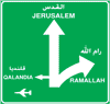 شاخصة مستطيلة كبيرة بأرضية خضراء وإطار أبيض، تحمل سهمًا أبيض متجهًا إلى الأعلى وسهمًا متفرعًا إلى اليمين وسهمًا إلى اليسار، وإلى جانب الأسهم أسماء «القدس JERUSALEM» و«رام الله RAMALLAH» و«قلنديا QALANDIA».